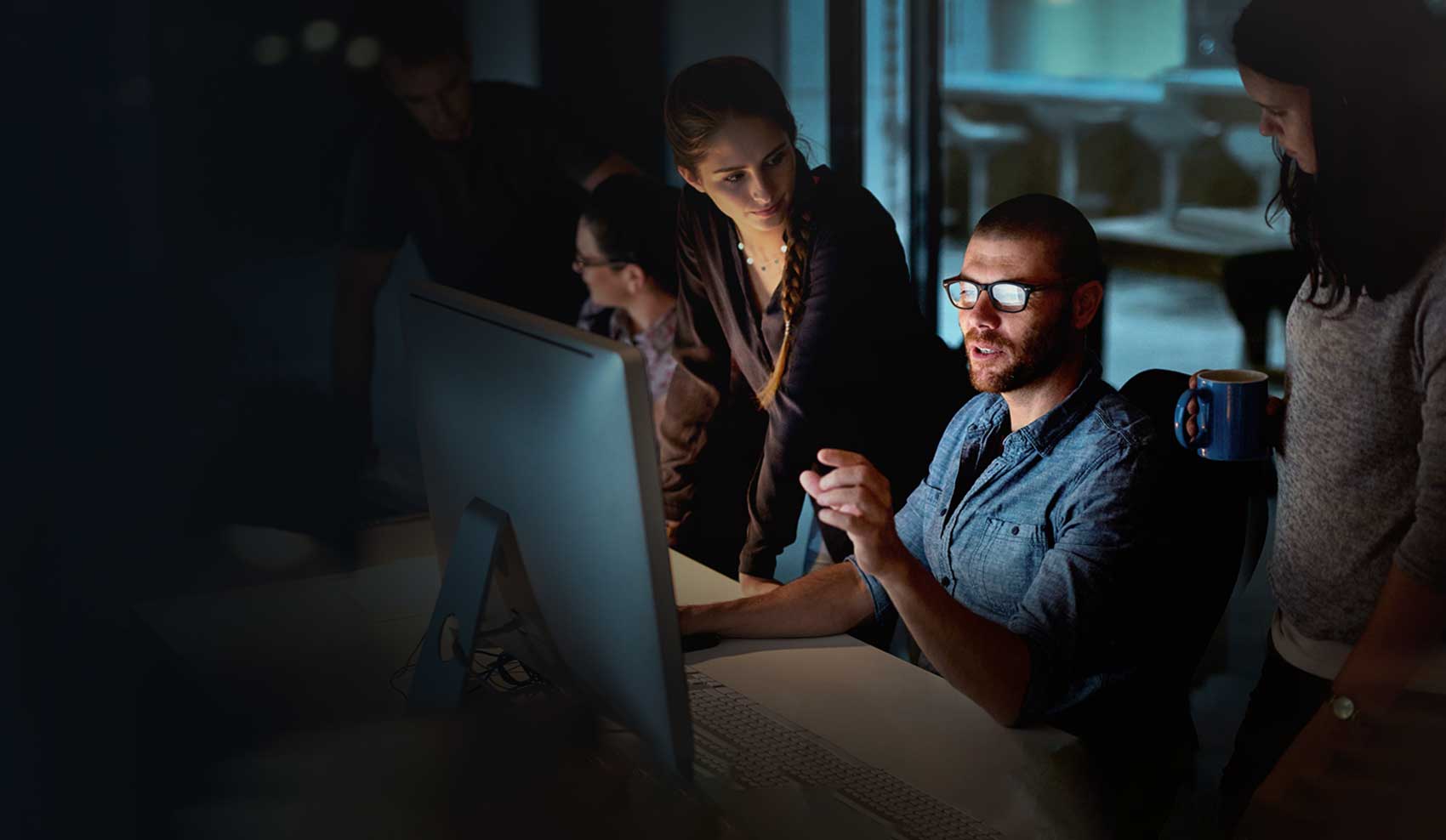 A group of professionals working in an office gathered around a large desktop monitor. A man in glasses is speaking while pointing at the screen, surrounded by three coworkers—two women and one man—engaged in discussion. The scene is dimly lit, suggesting a focused, collaborative environment.