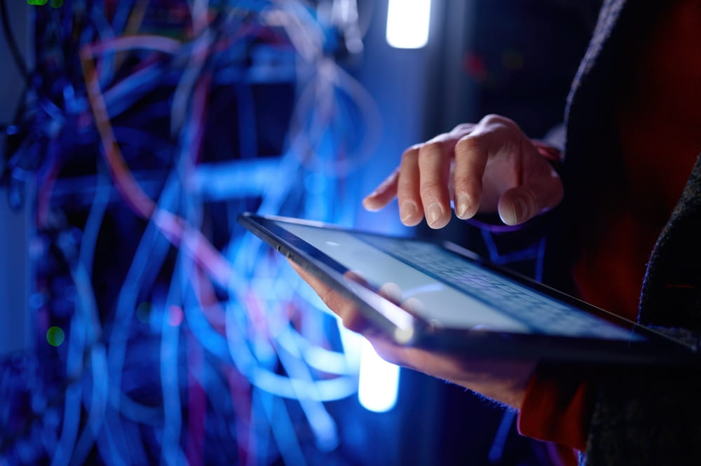 : A person uses a digital tablet next to a server rack with visible network cables